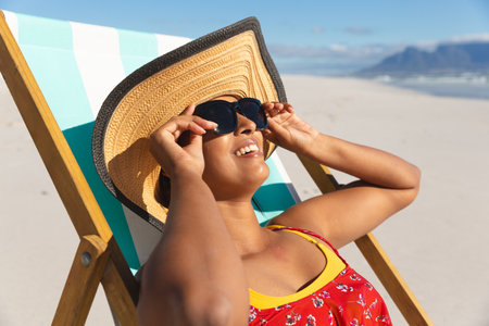 Smiling mixed race woman on beach holiday sitting in deckchair wearing sunglass sunbathing. healthy...