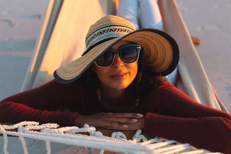Portrait of happy mixed race woman on beach holiday looking to camera lying in hammock during...