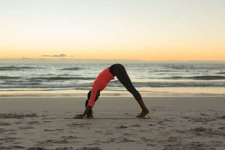 Mixed race woman on beach practicing yoga during sunset. healthy outdoor leisure time by the sea.の写真素材