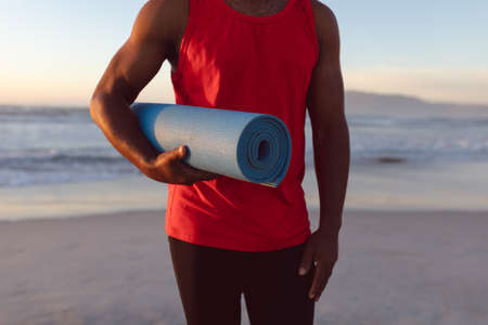 Mid section of senior african american man holding yoga mat at the beach. fitness yoga and healthy lifestyle conceptの写真素材