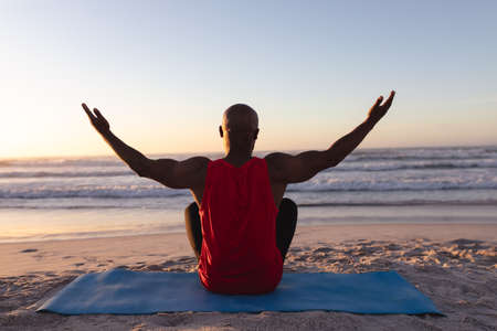 Rear view of senior african american man with arms wide open practicing yoga at the beach. fitness...