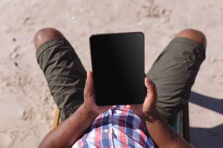 Overhead view of man holding digital tablet while sitting on deck chair at the beach. travel...