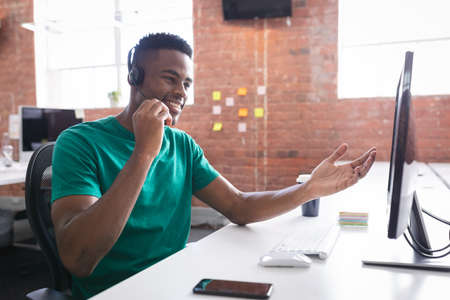 African american businessman having video call sitting in front of computer using headphones. independent creative design business.の写真素材