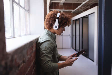 Portrait of happy mixed race businessman standing in office wearing headphones using tablet....