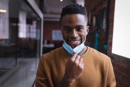 Portrait of happy african american businessman taking off face mask standing in corridor in office. independent creative design businessの写真素材