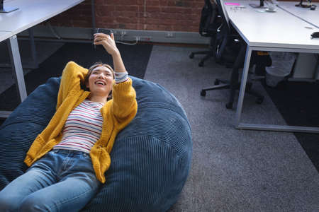 Happy mixed race businesswoman chilling in office relaxing space lying in beanbag using smartphone....