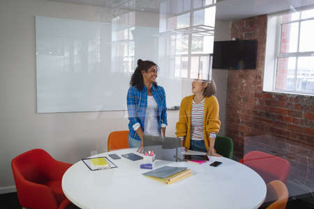 Happy diverse female colleagues standing at meeting room discussing over laptop. independent...
