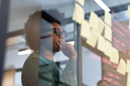 African american businesswoman looking at glass wall with sticky notes thinking. working person in a modern officeの写真素材