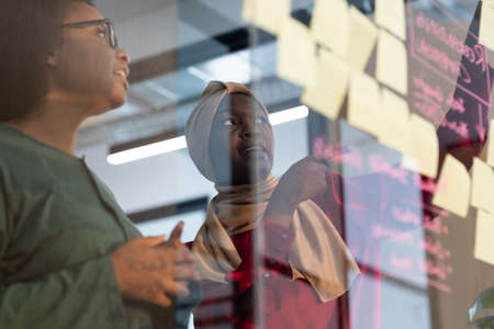 African american female business colleagues brainstorming putting sticky note on glass wall. working in a modern officeの写真素材