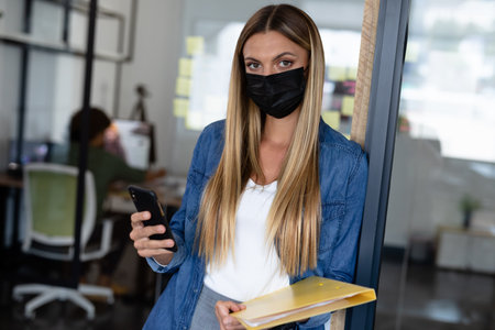 Caucasian businesswoman wearing mask looking to camera holding documents and smartphone. working in a modern officeの写真素材