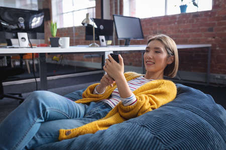 Happy mixed race businesswoman chilling in office relaxing space lying in beanbag using smartphone....