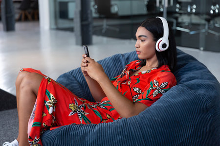 Portrait of mixed race businesswoman sitting in bean bag wearing headphones and using smartphone....