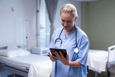 Smiling caucasian female doctor in hospital wearing scrubs and stethoscope using tablet. medical professional at work and health services.の写真素材