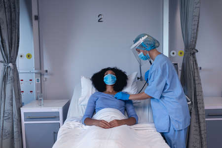 Caucasian female doctor in hospital wearing face mask examining female patient with stethoscope. medical professional at work during coronavirus covid 19 pandemic.の写真素材