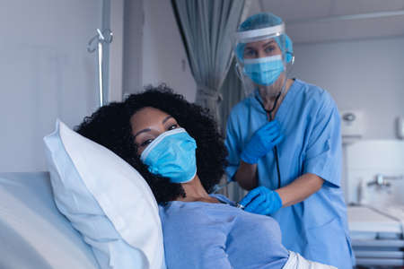 Caucasian female doctor in hospital in face mask examining female patient with stethoscope. medical professional at work during coronavirus covid 19 pandemic.の写真素材