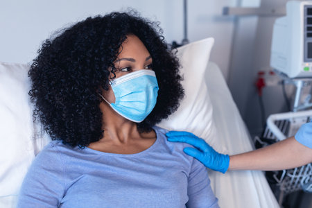 African american female patient in hospital bed wearing face mask consoled by female doctor. medical professional at work during coronavirus covid 19 pandemic.の写真素材