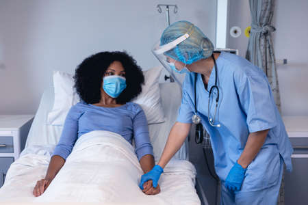 Caucasian female doctor in hospital in face mask consoling female patient in bed wearing face mask. medical professional at work during coronavirus covid 19 pandemic.の写真素材