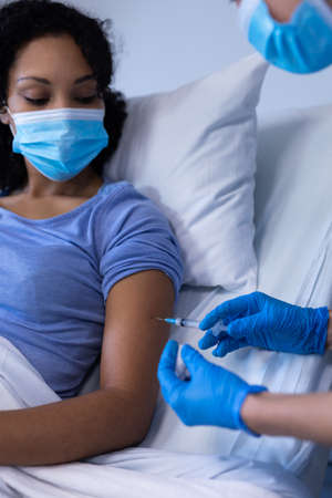 Caucasian female doctor in hospital wearing face mask vaccinating african american female patient....