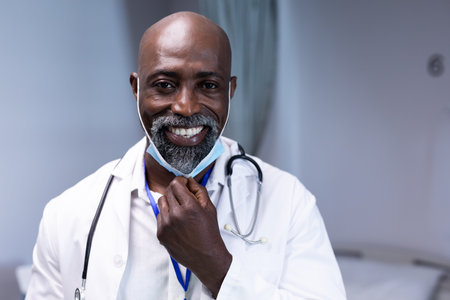 Portrait of african american male doctor in hospital smiling to camera with face mask. medical professional at work during coronavirus covid 19 pandemic.の写真素材