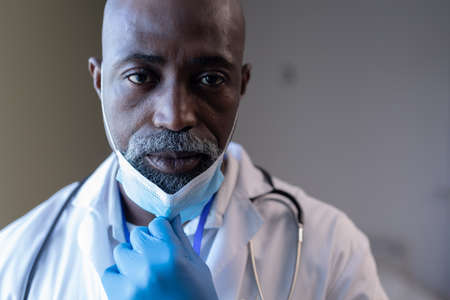 Portrait of worried african american male doctor in hospital holding face mask. medical professional at work during coronavirus covid 19 pandemic.の写真素材