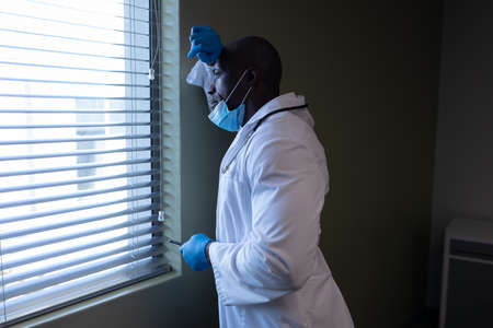 Thoughtful african american male doctor in hospital with face mask looking out of window. medical professional at work during coronavirus covid 19 pandemic.の写真素材
