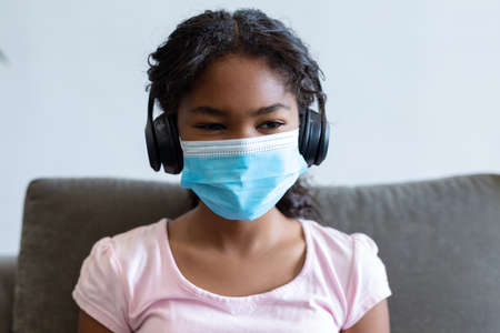 Portrait of mixed race girl wearing face mask and headphones. medicine and health during coronavirus covid 19 pandemic.の写真素材