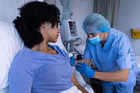Caucasian female doctor in hospital wearing face mask vaccinating african american female patient....