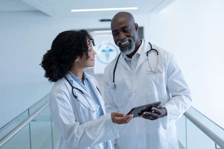 Smiling diverse male and female doctor talking and looking at digital tablet in hospital corridor. medicine, health and healthcare services.の写真素材