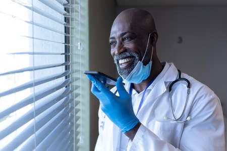 Smiling african american male doctor in hospital with face mask talking on smartphone. medical professional at work during coronavirus covid 19 pandemic.の写真素材