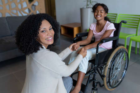 Happy mixed race mother kneeling holding hands with smiling daughter in wheelchair in hospital foyer. medicine, health and healthcare services.の写真素材