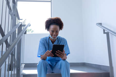 Smiling mixed race female doctor with face mask using tablet sitting on stairs in hospital. medical professional at work during coronavirus covid 19 pandemic.の写真素材