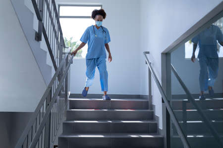 Mixed race female doctor wearing face mask and scrubs running down stairs in hospital. medical professional at work during coronavirus covid 19 pandemic.の写真素材