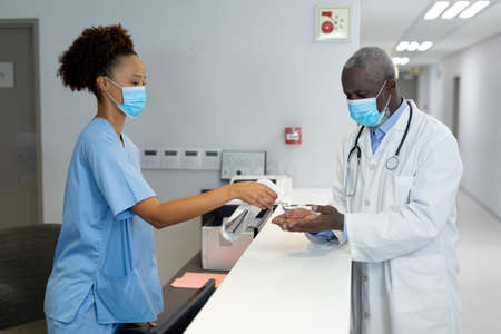 Diverse male and female doctors wearing face masks disinfecting hands over the counter at hospital. medicine, health and healthcare services during coronavirus covid 19 pandemic.の写真素材
