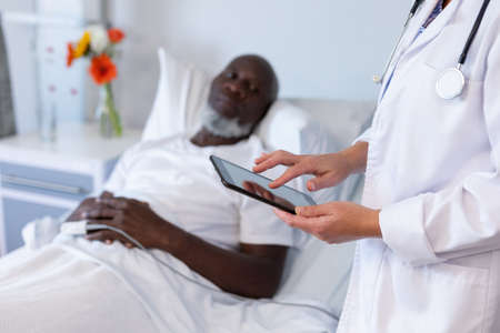 Caucasian female doctor standing next to african american male in hospital patient room using tablet. medicine, health and healthcare services.の写真素材