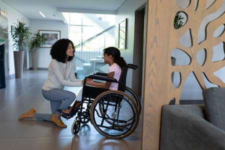 Happy mixed race mother kneeling holding hands talking with daughter in wheelchair in hospital foyer. medicine, health and healthcare services.の写真素材