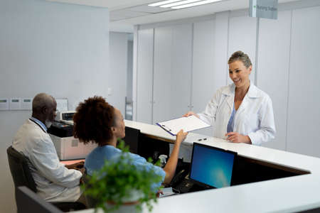 Caucasian female doctor passing document to african american receptionist at hospital reception. medicine, health and healthcare services.の写真素材