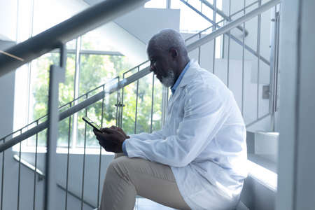 Senior african american male doctor using tablet sitting on stairs in hospital. medical professional at work.の写真素材