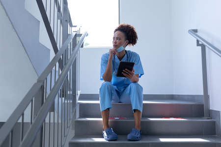 Mixed race female doctor with face mask holding tablet sitting on stairs in hospital. medical...