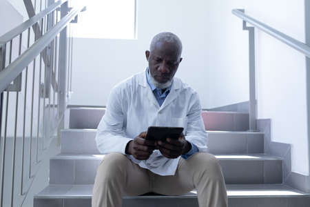 Senior african american male doctor using tablet sitting on stairs in hospital. medical professional at work.の写真素材