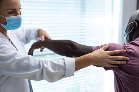 Caucasian female physiotherapist wearing mask stretching arm of african american male patient. medicine, health and healthcare services during coronavirus covid 19 pandemic.の写真素材
