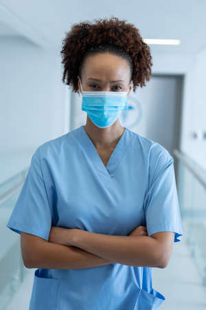 Portrait of african american female doctor wearing mask standing in hospital corridor. medicine, health and healthcare services during coronavirus covid 19 pandemic.の写真素材