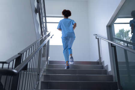 Rear view of mixed race female doctor wearing scrubs running up stairs in hospital. medical professional at work.の写真素材