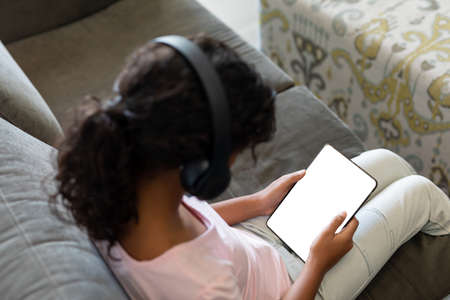Elevated view of mixed race girl wearing wireless headphones sitting on couch using digital tablet. teenage lifestyle, leisure time, communication and technology.の写真素材