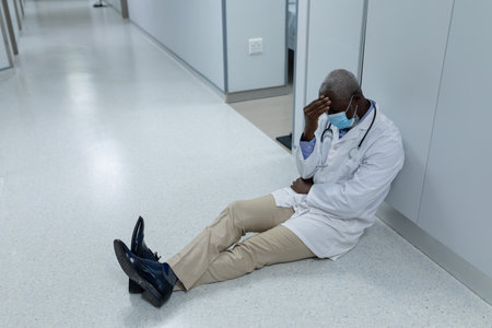 African american male doctor wearing mask sitting on floor in hospital corridor and worrying. medicine, health and healthcare services during coronavirus covid 19 pandemic. .の写真素材