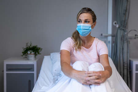 Portrait of caucasian female patient wearing mask sitting on hospital bed looking ahead. medicine, health and healthcare services during coronavirus covid 19 pandemic.の写真素材