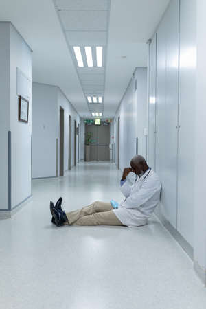 African american male doctor sitting on floor in hospital corridor and worrying. medicine, health and healthcare services.の写真素材