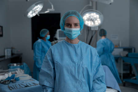 Portrait of caucasian female surgeon wearing face mask, surgical cap and gown in operating theatre. medicine, health and healthcare services during covid 19 coronavirus pandemic.の写真素材