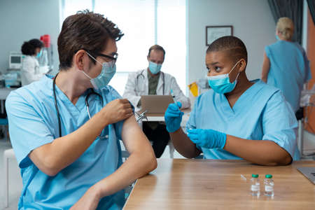 African american female doctor giving covid vaccination to male colleague, both in face masks....
