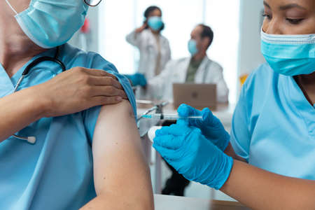 African american female doctor giving covid vaccination to male colleague, both in face masks. medicine, health and healthcare services during covid 19 coronavirus pandemic.の写真素材