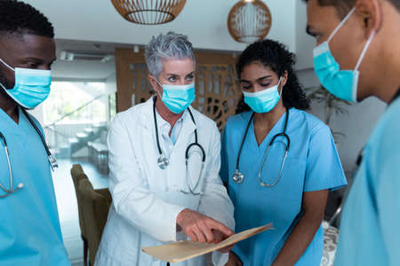Caucasian female senior doctor and diverse hospital colleagues wearing face masks talking at meeting. medicine, health and healthcare services during covid 19 coronavirus pandemic.の写真素材